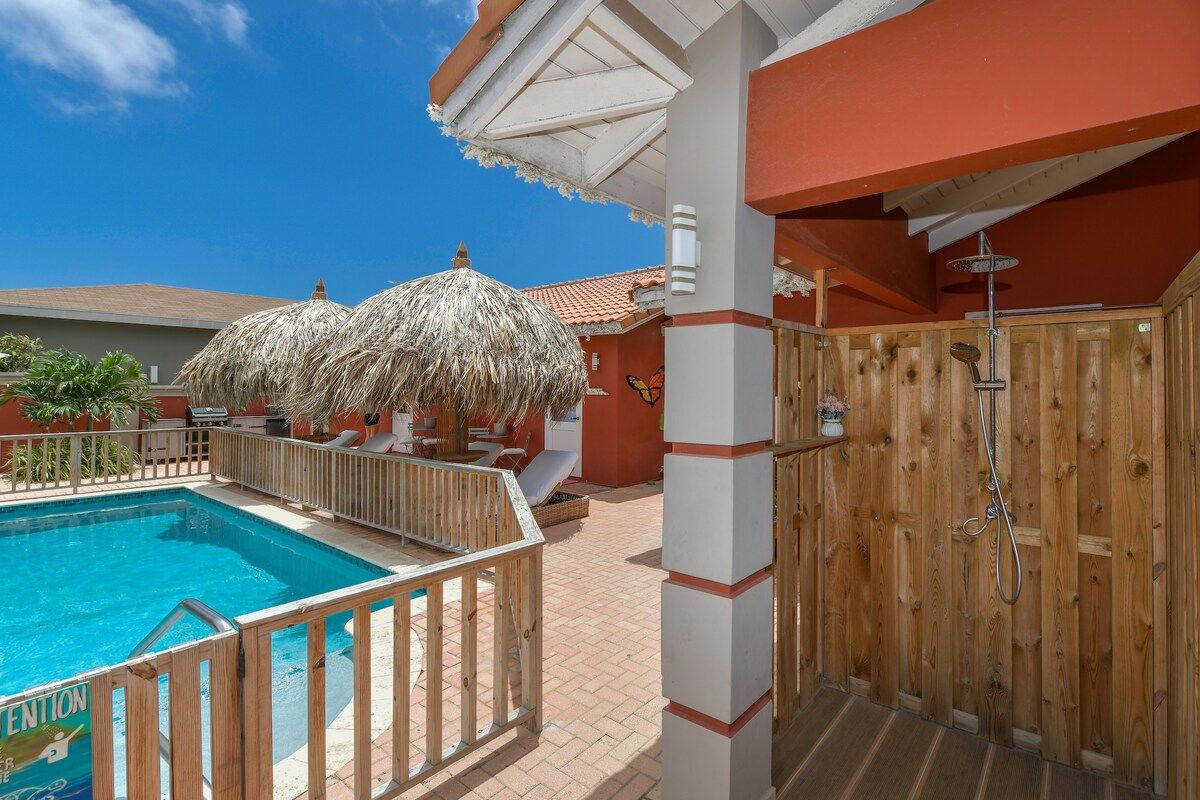 A swimming pool with a wooden deck, a wooden shower enclosure, and thatched umbrellas.