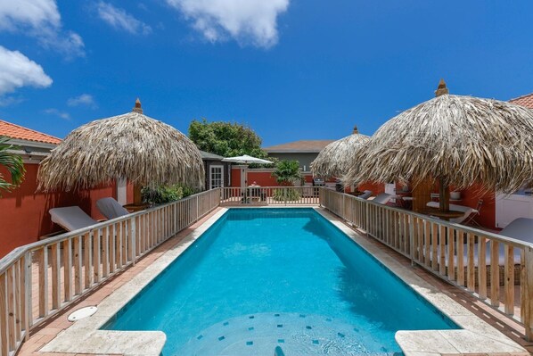 Una piscina rectangular con una terraza de madera y sombrillas de paja, rodeada de sillas de descanso y una valla de madera.