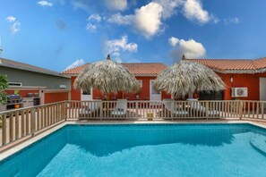 Una piscina con muelle de madera y sombrillas de paja, rodeada de una zona vallada con un edificio rojo al fondo.