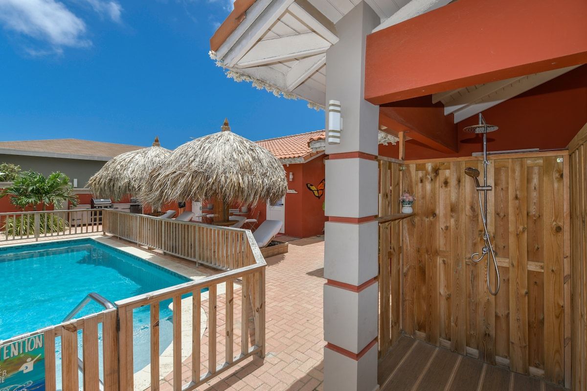 A pool area with a wooden deck, a covered seating area, and a wooden wall with a shower.