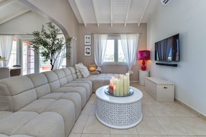 A living room with a sectional sofa, a round coffee table with candles, a television on the wall, and a large window with curtains. - Casa Lou Lou Beachhouse 2bdr 2Bth shared pool - central location (Tanki Leendert)