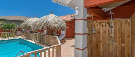 A pool area with a wooden deck, a covered seating area, and a wooden wall with a shower.