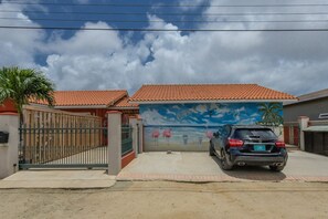 A gated residential area with a mural of flamingos and palm trees on the wall, a parked car, and a clear sky. - Studio Palm by Casa Lou Lou- modern  with shared pool- central location (Noord)