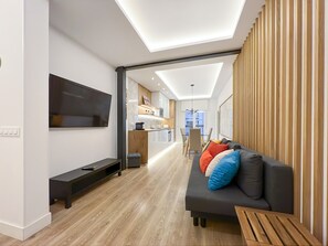 Una sala moderna con un televisor de pantalla plana, un sofá con cojines de colores, una mesa de centro de madera y una zona de cocina al fondo.