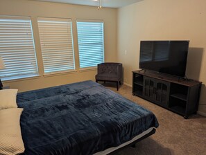 Un dormitorio con una cama, un televisor sobre un mueble, una silla y tres ventanas con persianas.