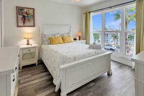 A bedroom with a bed, nightstands, a window with curtains, and a painting on the wall. - Madeira Beach Yacht Club Condo with Heated Pool, Marina, Beautiful Grounds and Easy Beach Access (Madeira Beach)