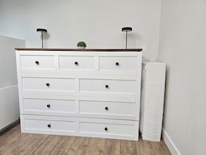Un aparador blanco con varios cajones y dos lámparas negras encima, ubicado en una habitación con piso de madera y pared blanca.