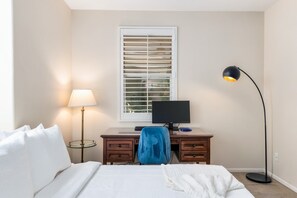 A bedroom with a bed, a desk with a computer, a lamp, and a window with blinds. - For Shore | Private Pool & Spa | Fire pit | Rancho Mirage (Rancho Mirage)
