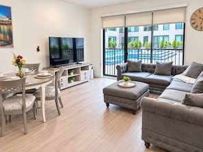A modern living room with a sofa, ottoman, dining table, chairs, a television, and a view of a swimming pool. - Downtown Cincinnati | Free Parking, Pool & Gym (Cincinnati)