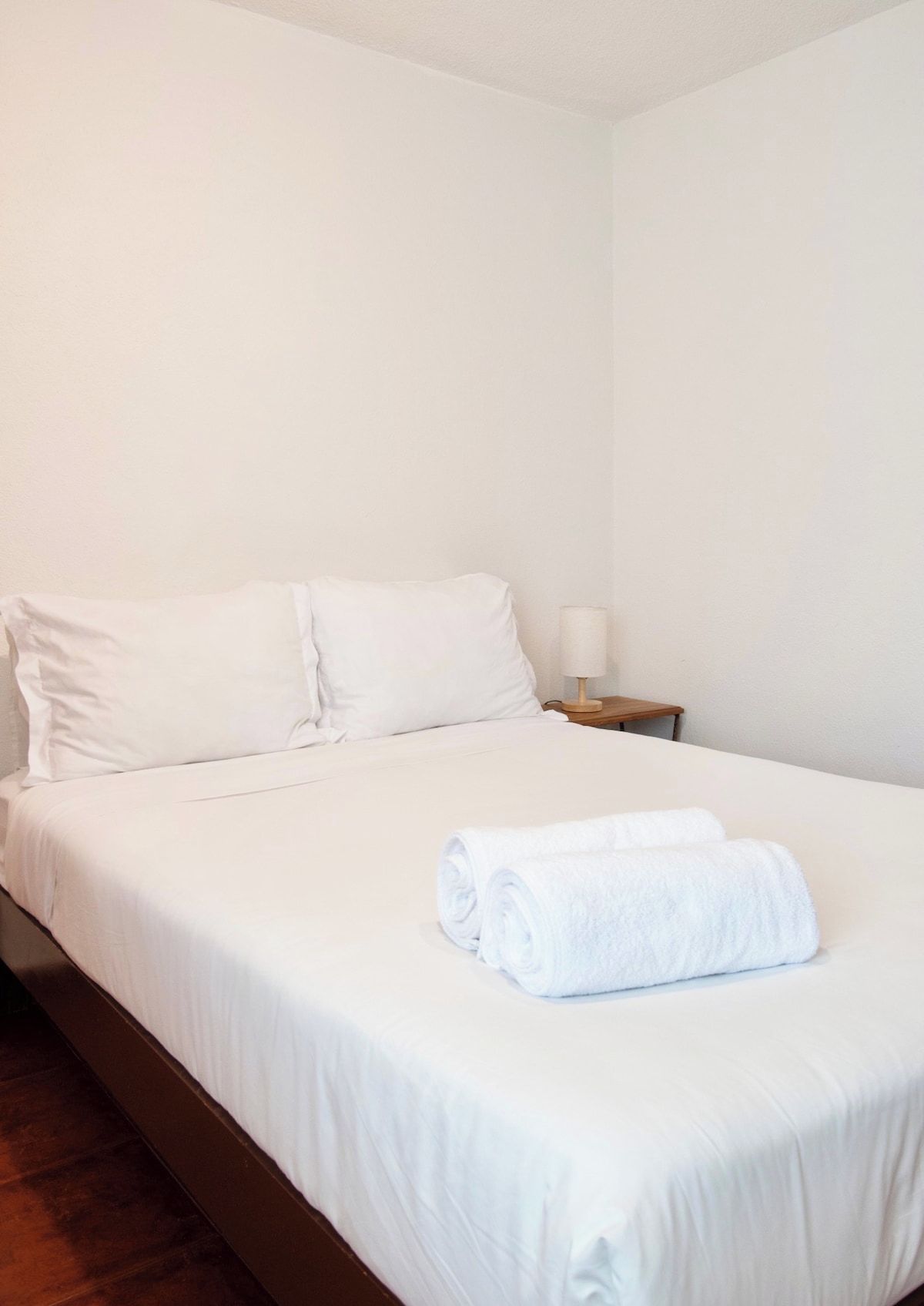 A single bed with white bedding, a wooden nightstand with a lamp, and a rolled white towel on the bed.