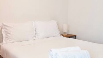 A single bed with white bedding, a wooden nightstand with a lamp, and a rolled white towel on the bed.