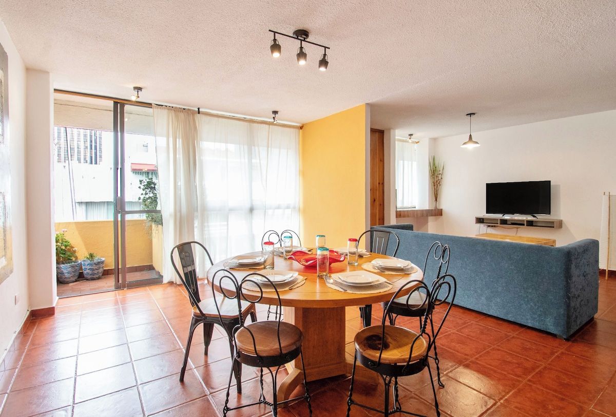 A dining area with a round table set for six, surrounded by metal chairs, a blue sofa, and a television on a stand.