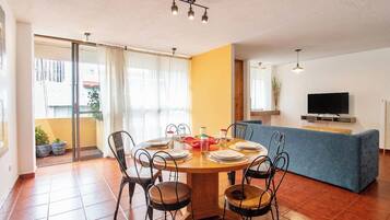 A dining area with a round table set for six, surrounded by metal chairs, a blue sofa, and a television on a stand.