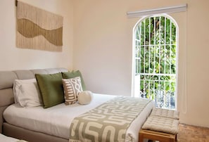 A bedroom with a bed, pillows, a bench, a window with a view of greenery, and a wall-mounted artwork. - Casa Raíz| A Place to Belong, Rooted in Connection (Mexico City)
