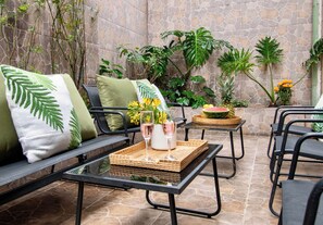 Un espace patio comprenant une table avec un plateau en verre, deux chaises, un canapé avec des coussins verts, ainsi que diverses plantes et fleurs.