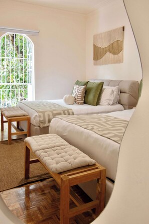 A bedroom with two beds, a wooden bench, and a view of greenery through a window with a white trellis. - Casa Raíz| A Place to Belong, Rooted in Connection (Mexico City)