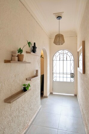 Un couloir aux murs blancs à texture, des étagères en bois ornées d’objets décoratifs, un luminaire suspendu et une grande fenêtre en arc surmontée d’un motif en métal.