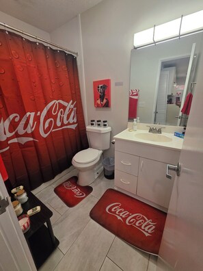 Un baño con inodoro, lavamanos y cortina de ducha con el logotipo de Coca-Cola. Hay dos botellas sobre la encimera del lavamanos.
