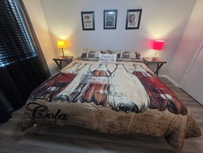 Un dormitorio con una cama que presenta un diseño vintage de Coca-Cola, dos mesitas de noche con lámparas y cuadros enmarcados en la pared.