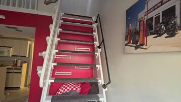 Una escalera con peldaños rojos y barandilla blanca, que lleva a una habitación con cocina al fondo.