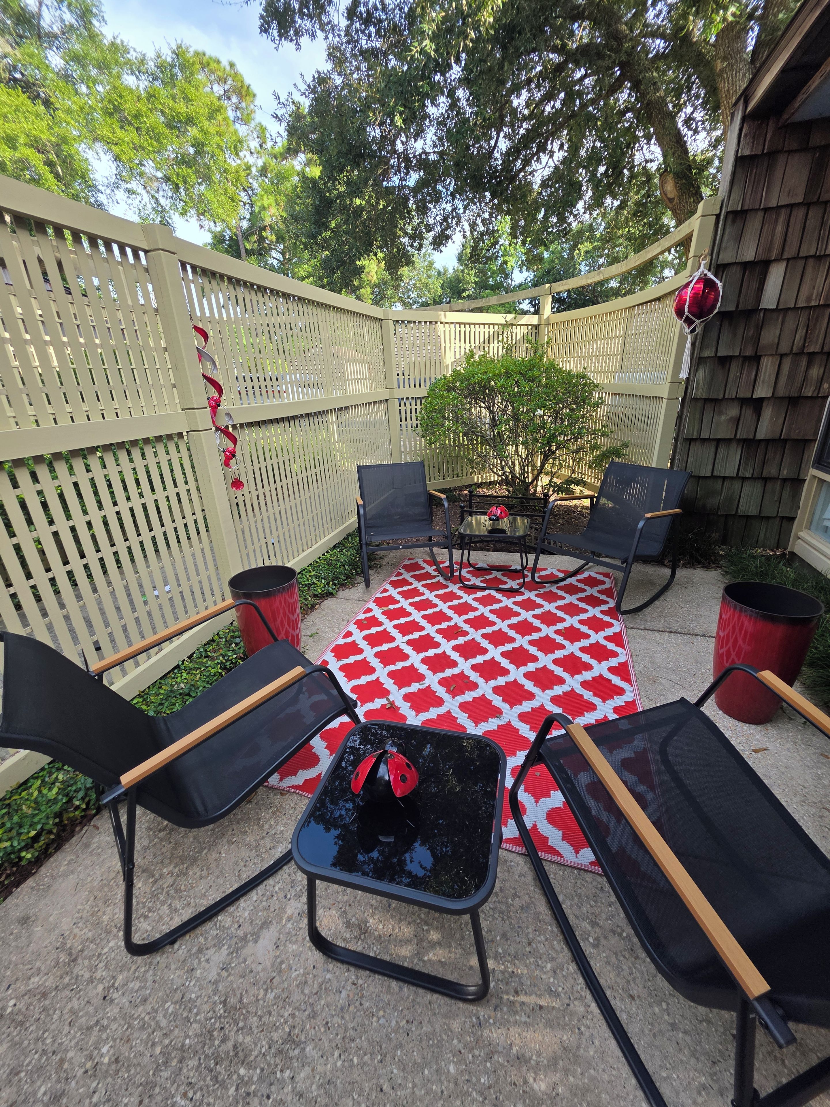 Un patio al aire libre con un mantel rojo y blanco estampado, sillas negras y una mesita con un objeto rojo encima.