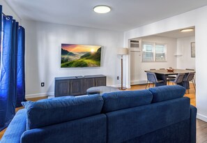 A modern living room with a blue sofa, a dining table with chairs, and a painting of a landscape on the wall. - Jersey City Heights 3BR condo, sleeps 6, private entrance + driveway parking (Jersey City)