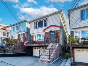 A residential street with houses featuring front porches, a garage, and a visible American flag. - Jersey City Heights 3BR condo, sleeps 6, private entrance + driveway parking (Jersey City)