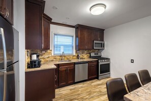 A kitchen with dark wooden cabinets, a stainless steel refrigerator, a microwave, a stove, and a dishwasher. There is a window with blinds, a sink, and a countertop with a coffee maker and toaster. - Jersey City Heights 3BR condo, sleeps 6, private entrance + driveway parking (Jersey City)