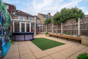 A backyard with a hot tub, artificial grass, a wooden bench, and a wooden fence.
