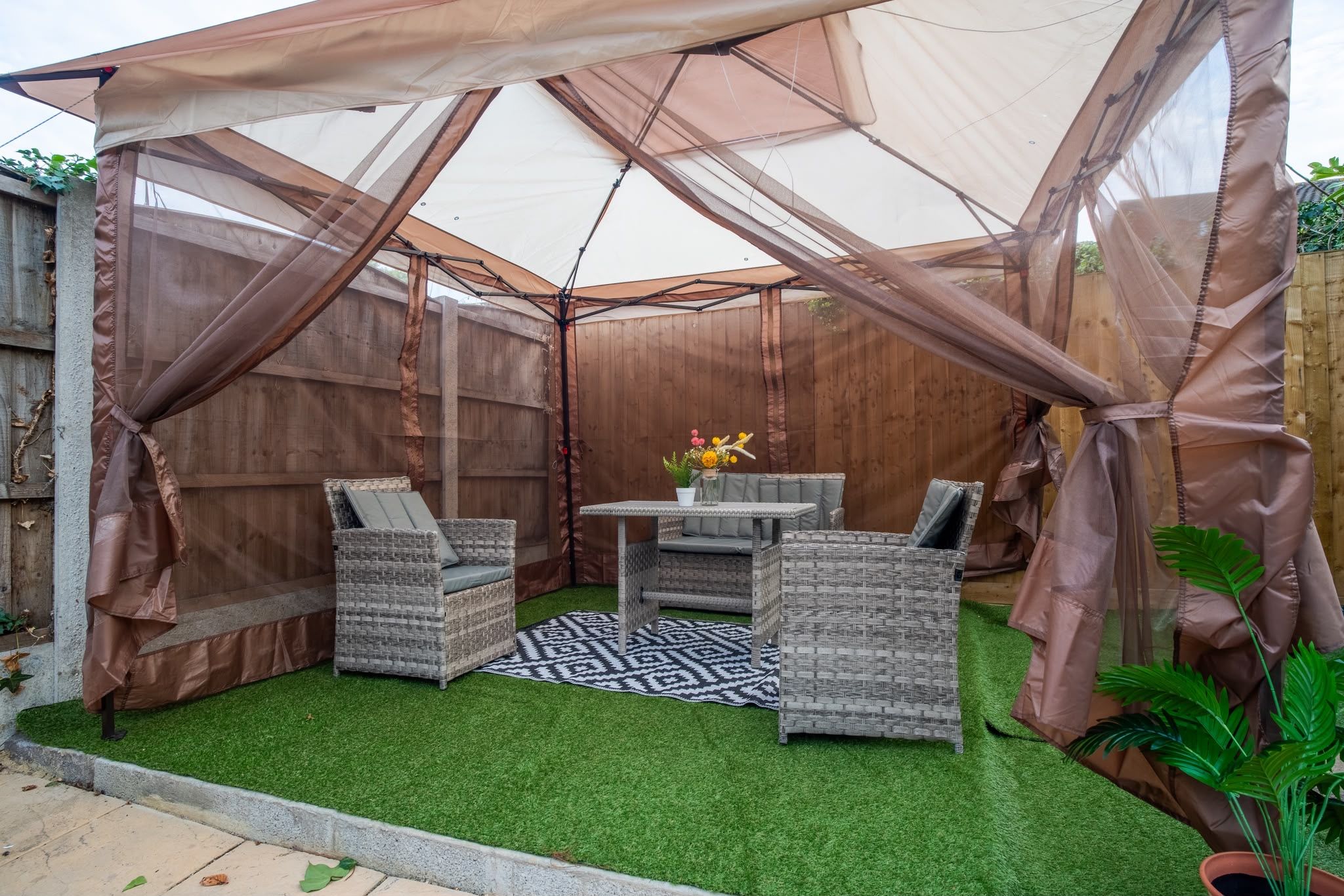 A covered outdoor seating area with wicker furniture and a central table, surrounded by a wooden fence and a white canopy.