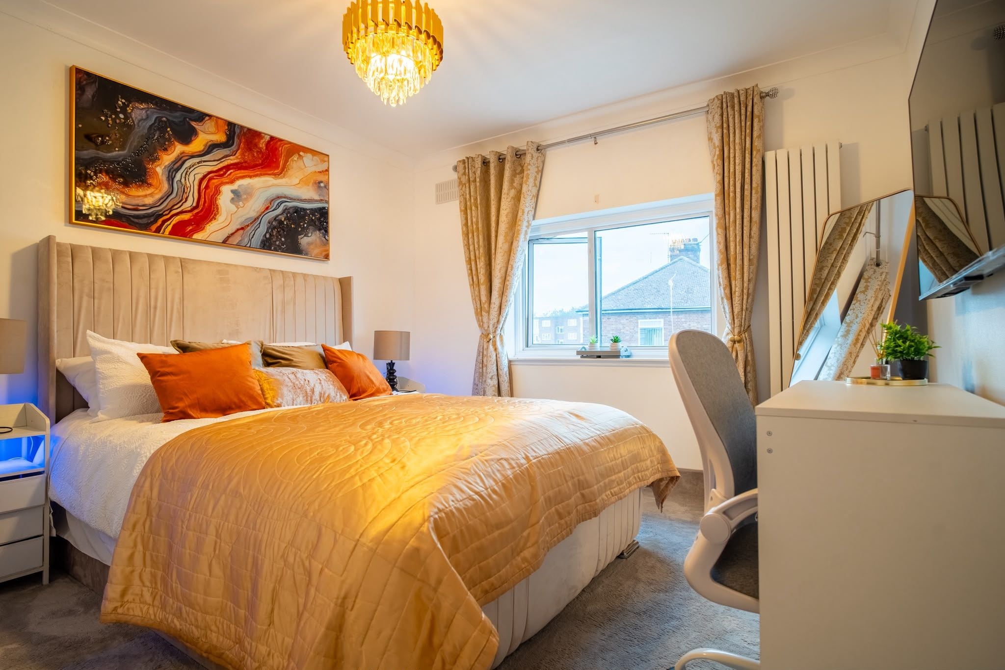 A hotel room with a bed, a desk, a chair, a window with curtains, a painting on the wall, and a chandelier.