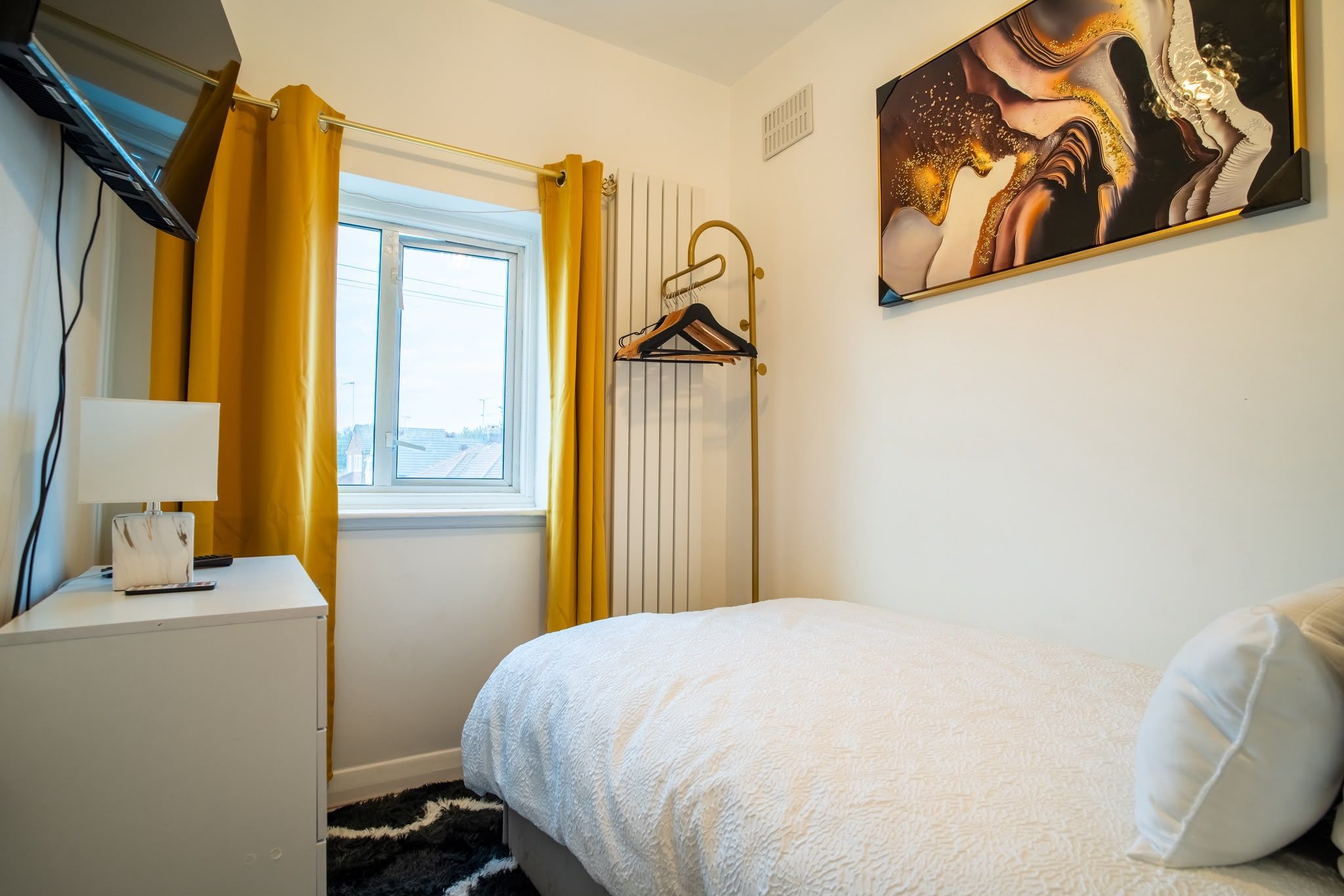 A bedroom with a bed, a bedside table, a lamp, a window with curtains, a radiator, and a painting on the wall.