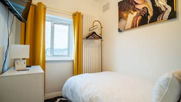 A bedroom with a bed, a bedside table, a lamp, a window with curtains, a radiator, and a painting on the wall.