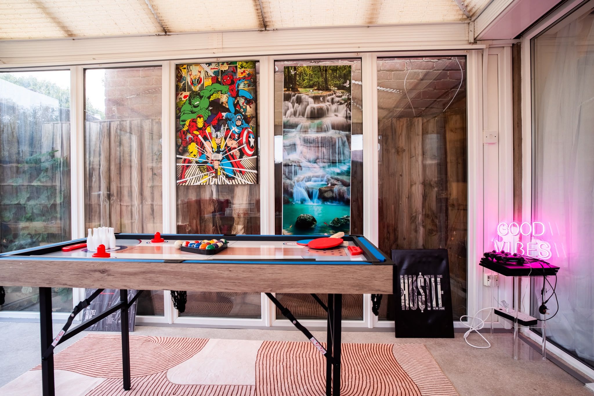 A pool table in a room with large windows, a neon sign, and a poster of comic book characters.