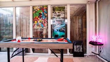 A pool table in a room with large windows, a neon sign, and a poster of comic book characters.