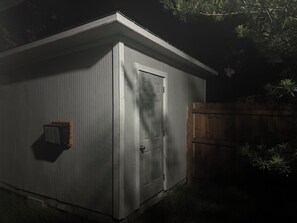 A metal shed with a door and a small window, surrounded by a wooden fence and some vegetation.