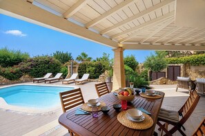 Een buitenzwembad met ligbedden, een eettafel met kommen en glazen, en een pergola erboven.