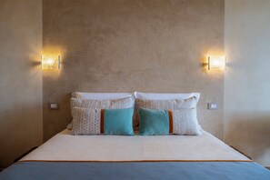 A bed with a white mattress and two pillows, one with a pattern and one solid teal, against a textured wall with two wall-mounted lights. - Villa Tigli: A 5-Minute Walk From the Beach by Top Villas Sardinia (Quartu Sant'Elena)