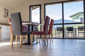 Spiseområde med et bord og stole, store vinduer med udsigt til et bjerglandskab, samt et køkken med skabe og en bordplade.