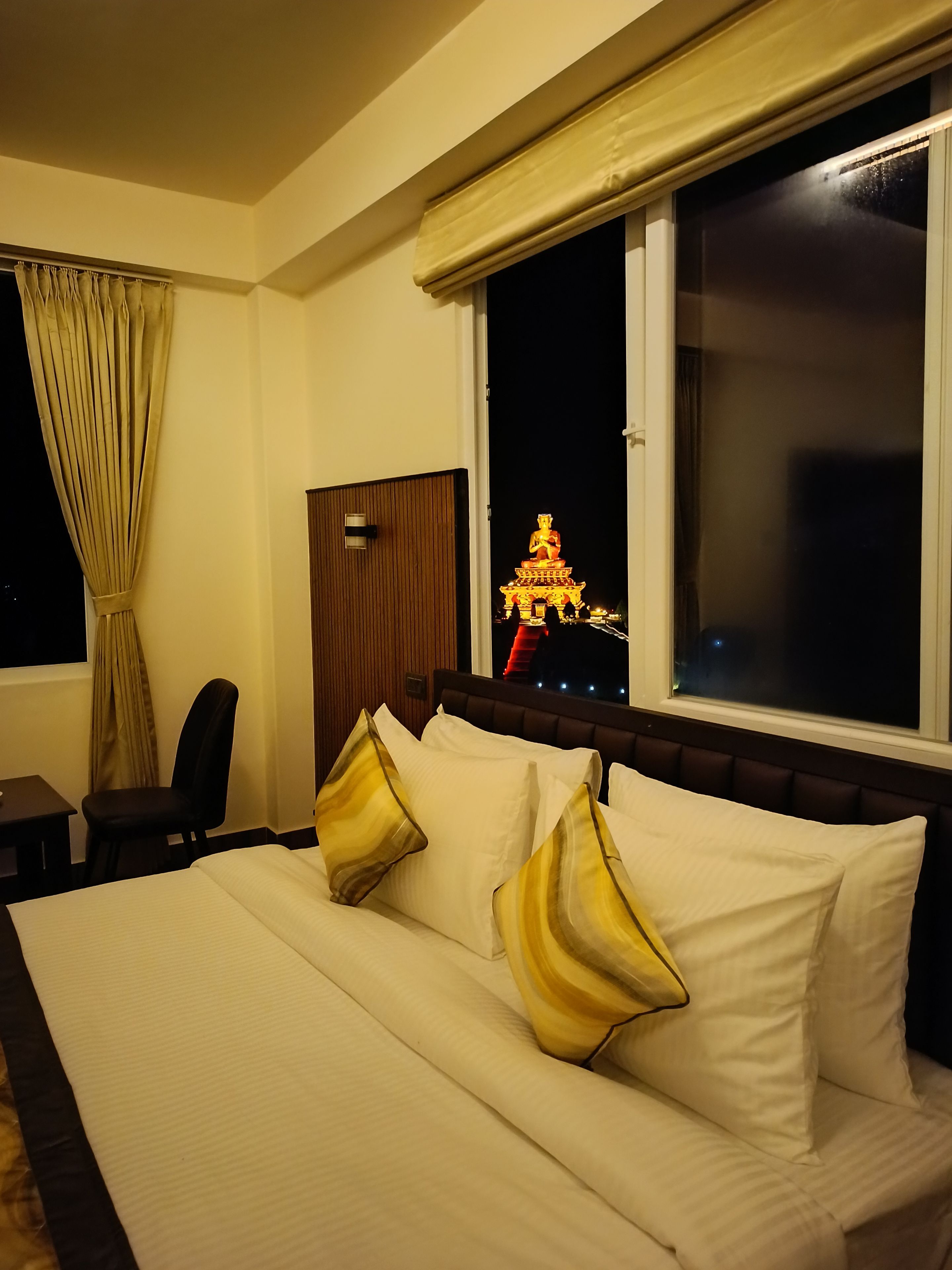 A hotel room with a bed, a chair, a desk, and a window with a view of a lit temple at night.