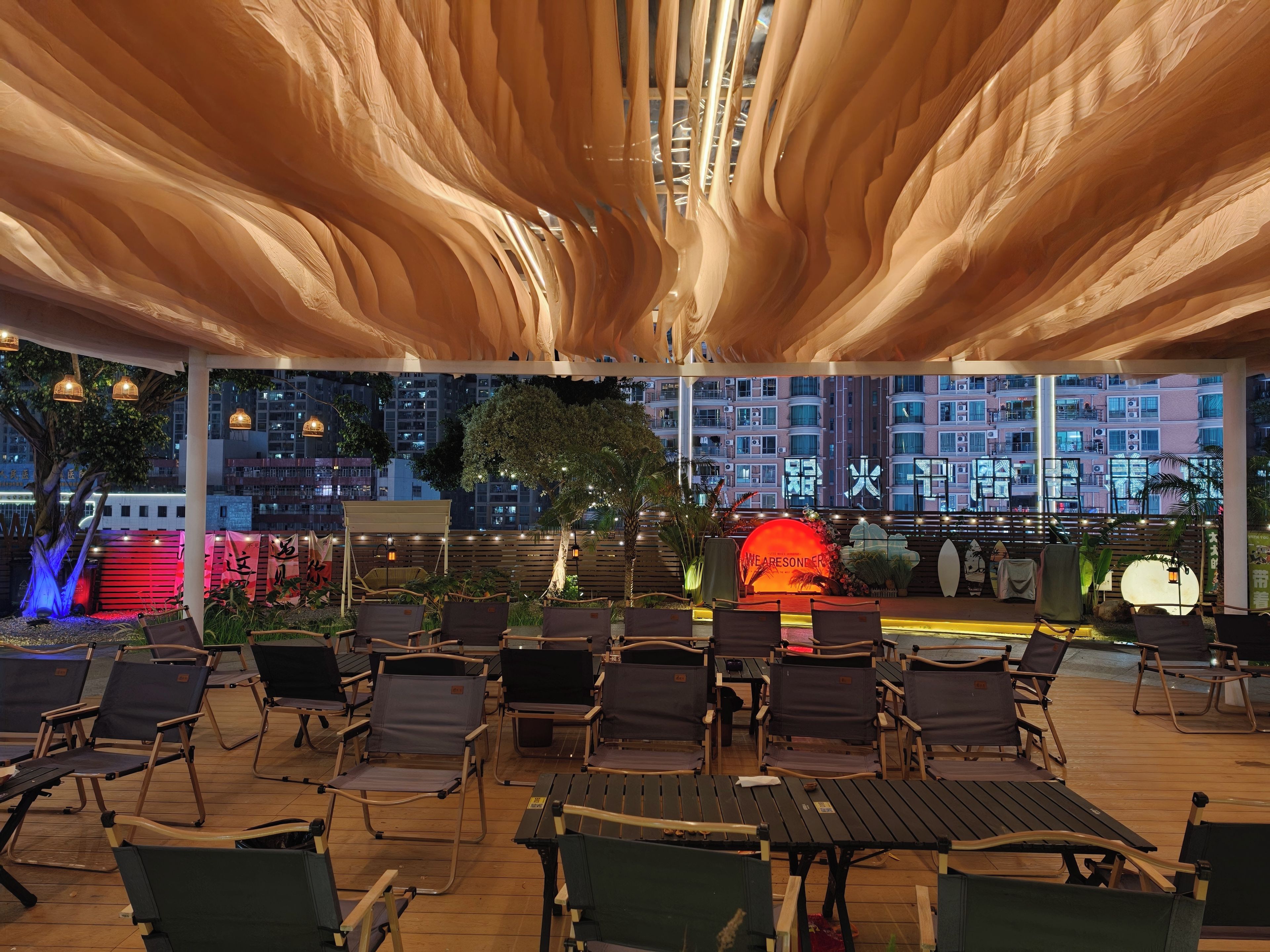 An outdoor seating area with chairs and tables, a decorative wall with illuminated text, and a view of a cityscape at night.
