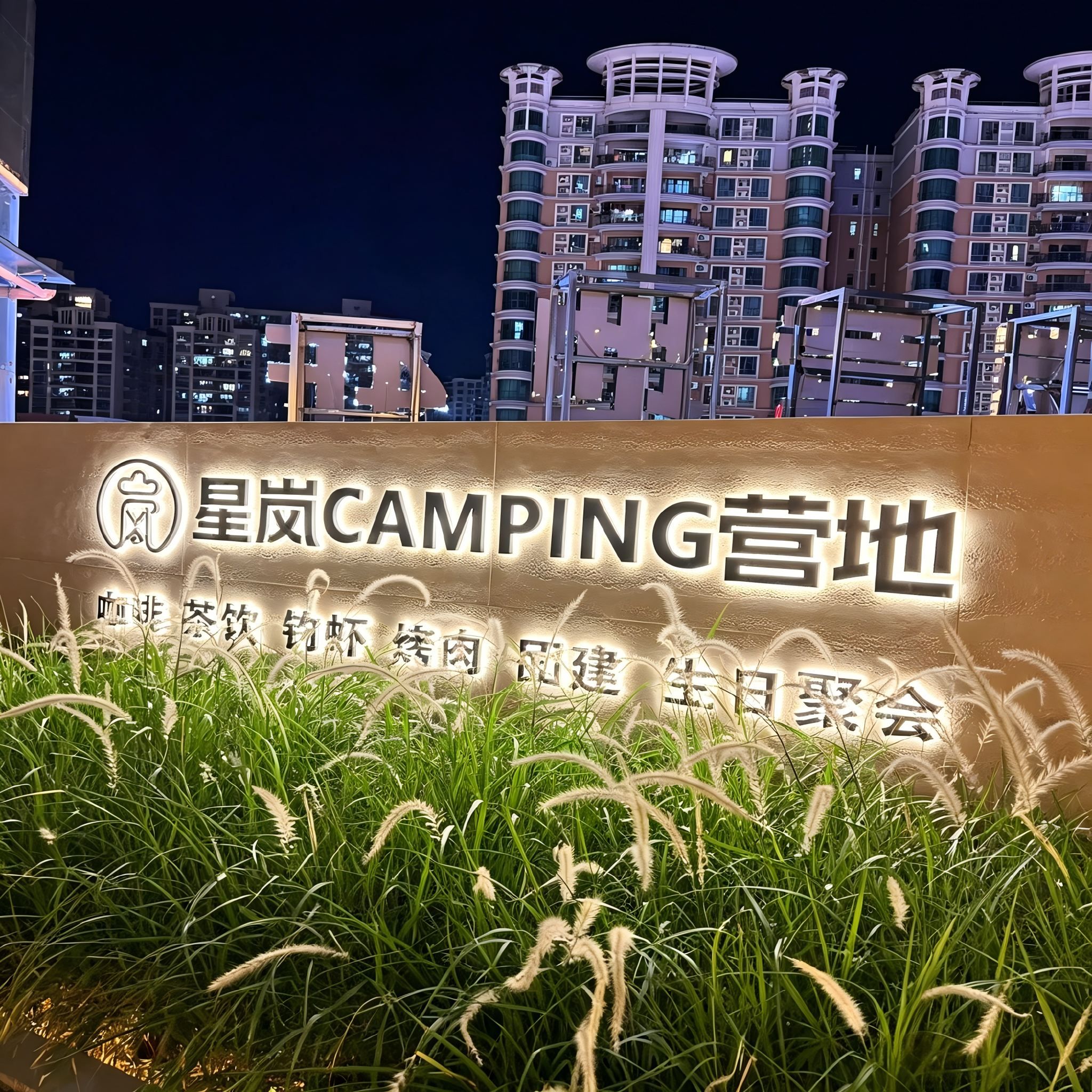 Un letrero que dice '星岗CAMPING营地' con vistas a edificios modernos al fondo.
