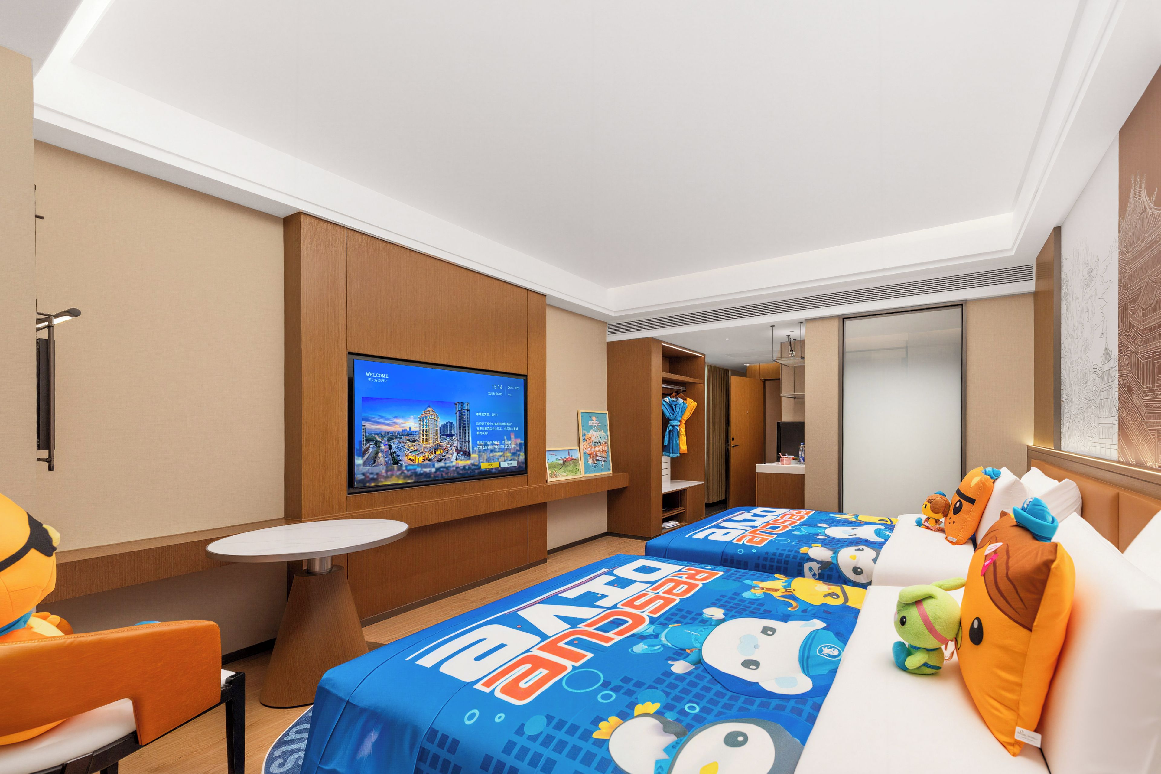 A hotel room with a large TV, two beds with cartoon-themed bedding, a small table, and a seating area.