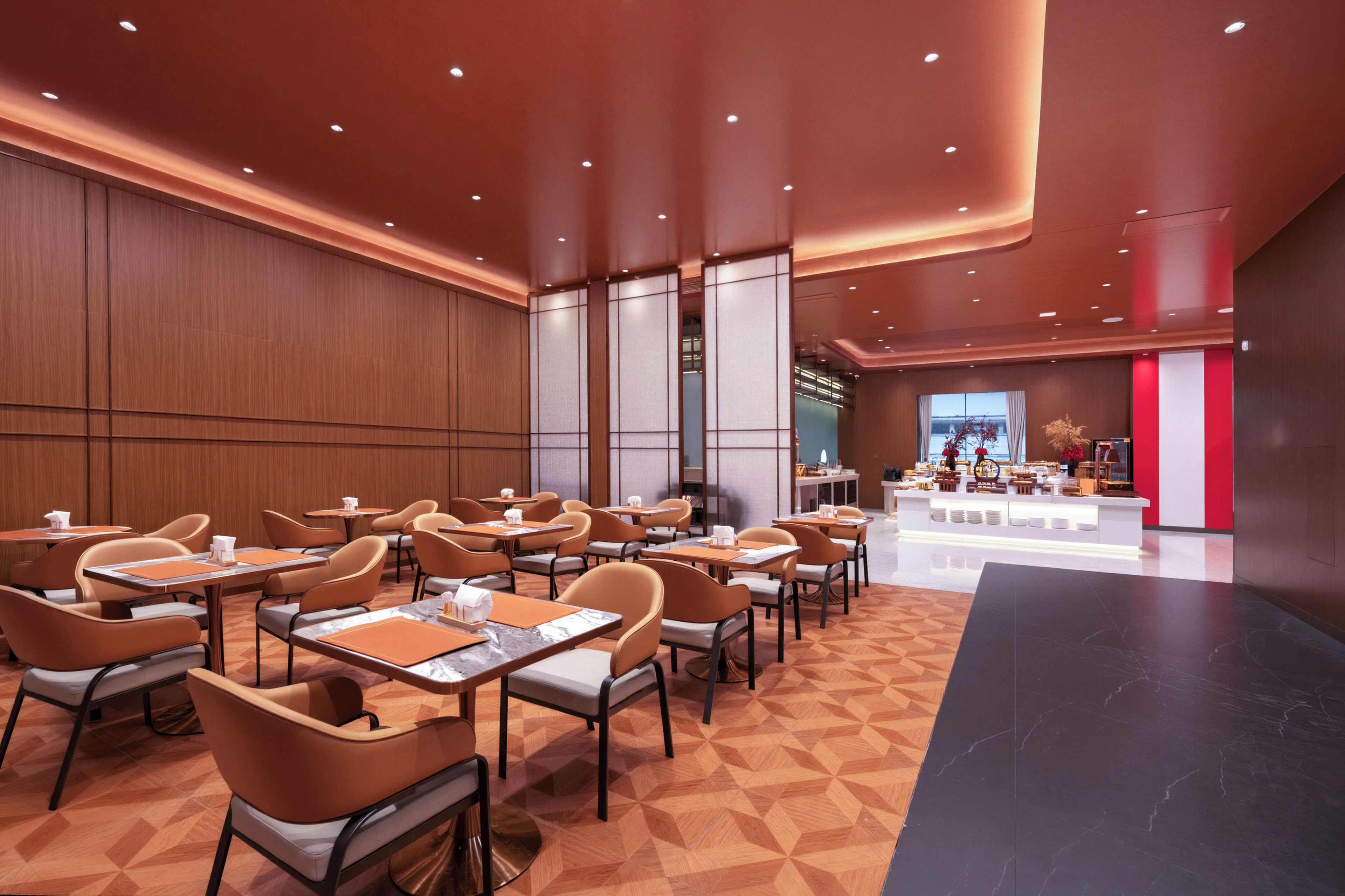 A modern hotel lobby with a long reception desk, multiple seating areas with tables and chairs, and a large window offering a view.