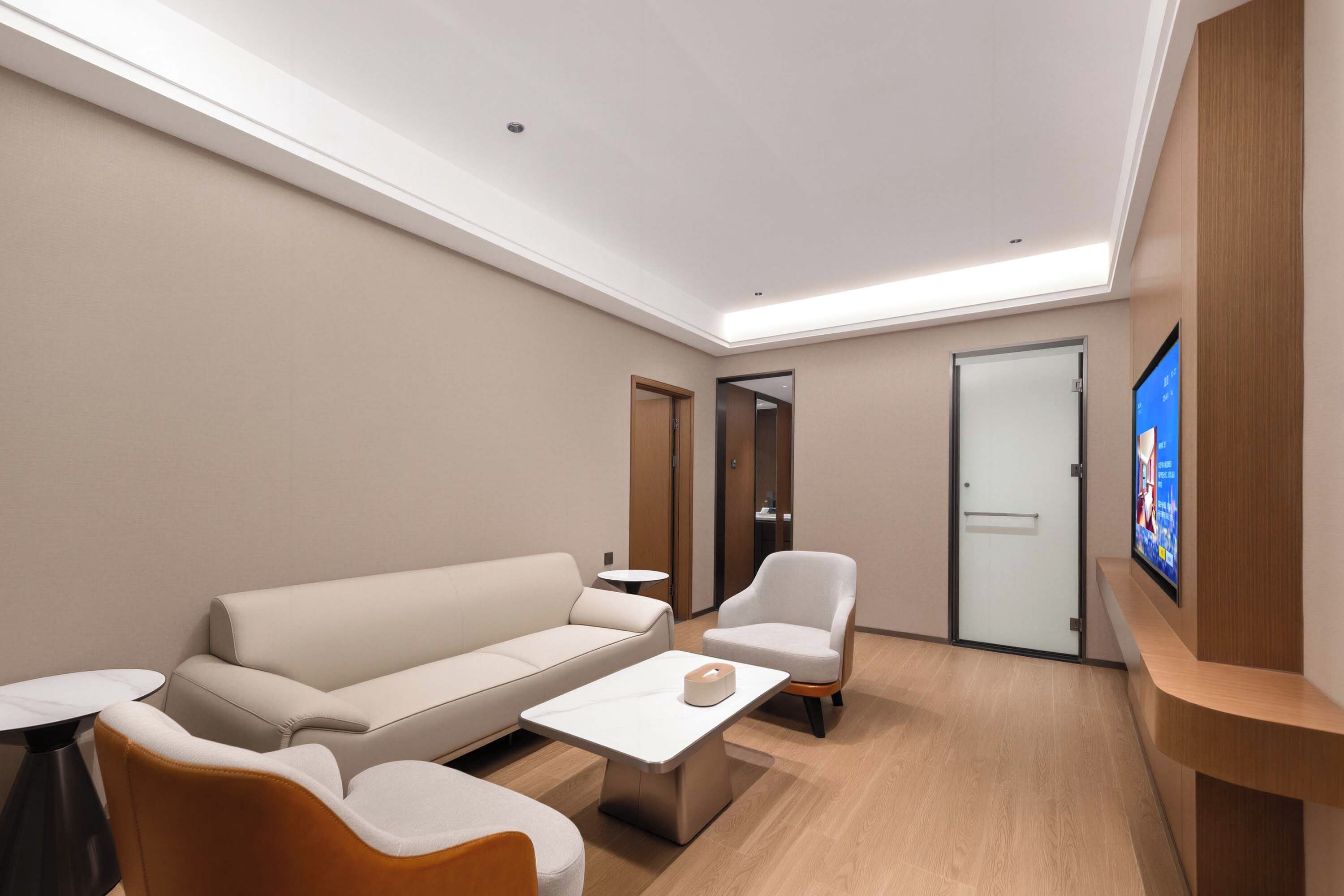 A modern hotel room with a sofa, armchair, coffee table, and a flat-screen TV mounted on the wall.