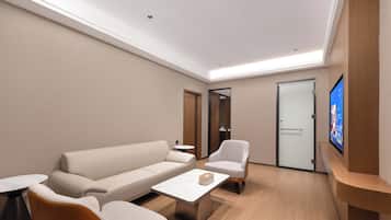 A modern hotel room with a sofa, armchair, coffee table, and a flat-screen TV mounted on the wall.
