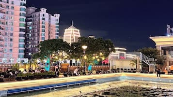 A nighttime view of a cityscape with a large illuminated building in the background, a water feature with lily pads in the foreground, and a seating area with people.