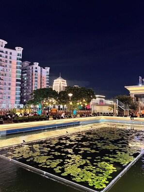 A nighttime view of a cityscape with a large illuminated building in the background, a water feature with lily pads in the foreground, and a seating area with people. - Haofeng Wyndham Zhongshan Guzhen Guomao (Zhongshan)