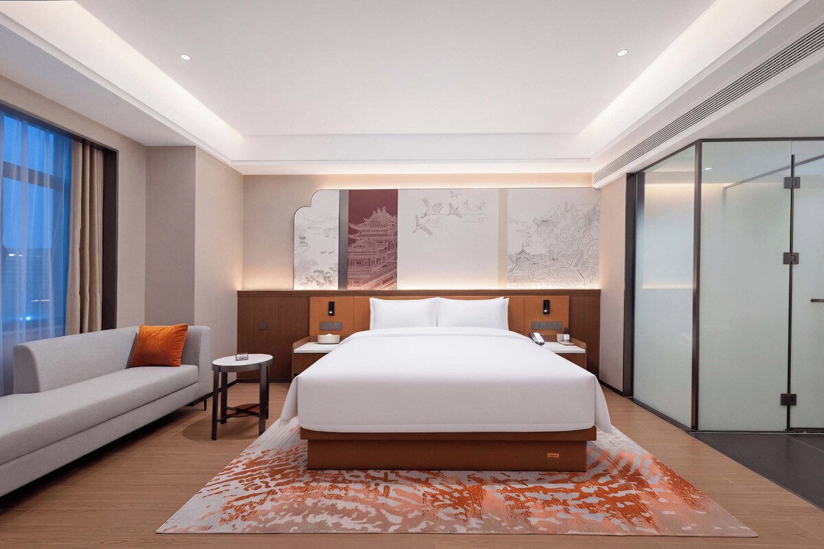 Une chambre d’hôtel moderne avec un grand lit, un canapé, une petite table et une cloison en verre.