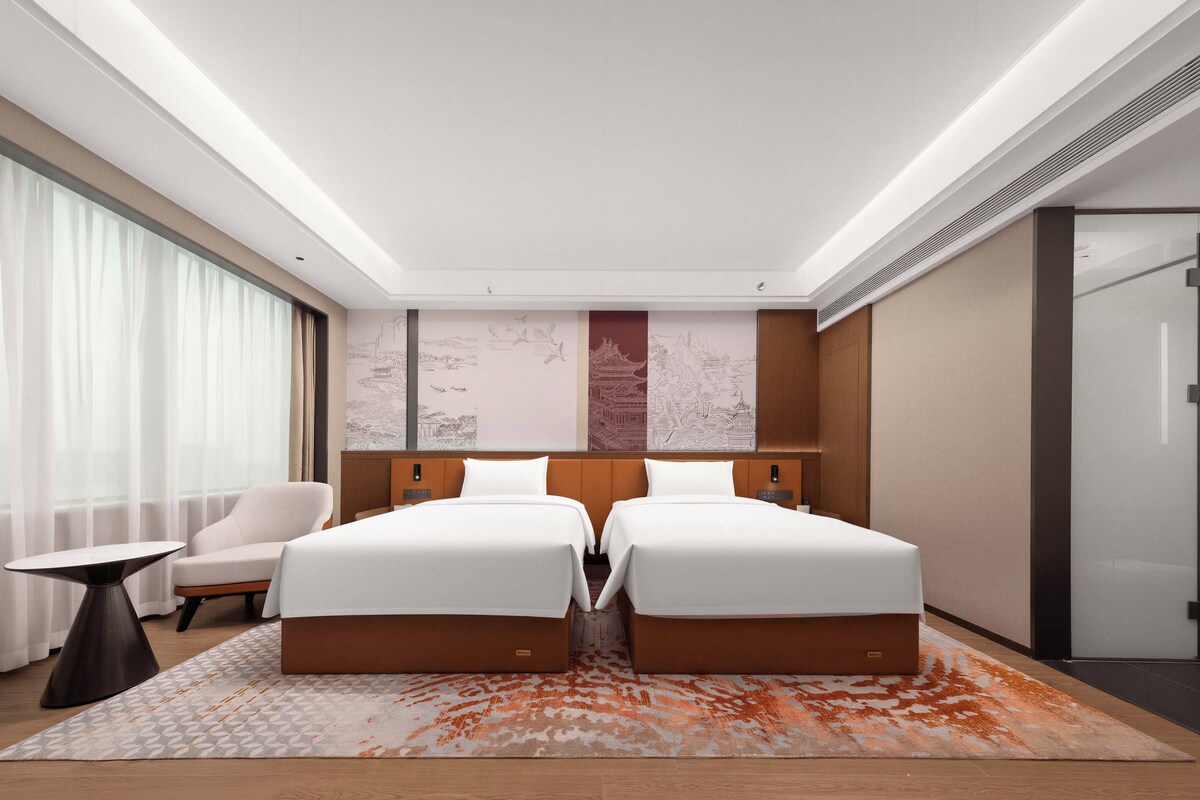 Une chambre d’hôtel moderne avec un grand lit, une chaise, une petite table et une grande fenêtre avec des rideaux.